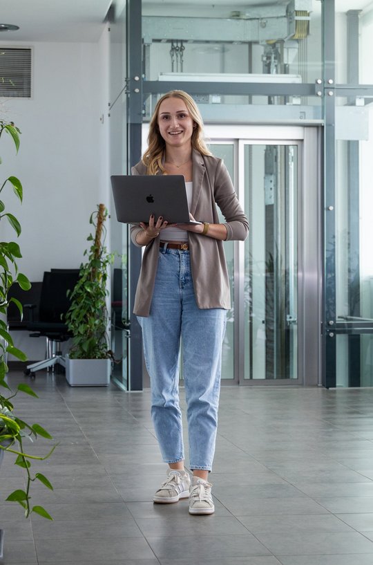 Die junge Frau in Jeans und Blazer strahlt Vertrauen aus, während sie ihren Laptop in einem lichtdurchfluteten Bürobereich hält.