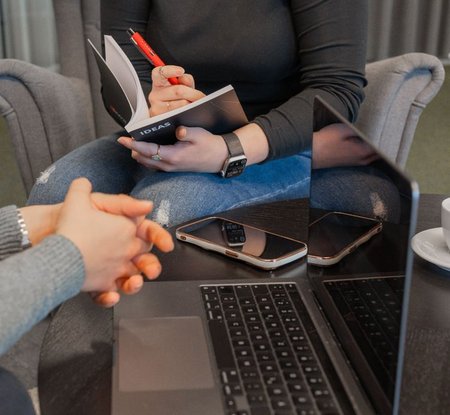 Ausschnitt der Hände von zwei Mitarbeiterinnen mit Buch und Stift in der Hand und vor dem Laptop