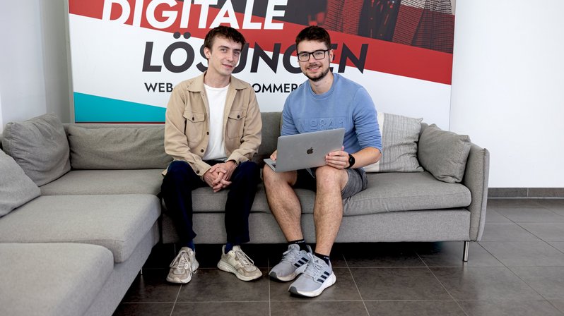Zwei Männer lächeln freundlich und sitzen gemeinsam auf einer grauen Couch mit dem Laptop